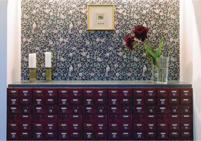 a room with a wallpapered wall, a wooden cabinet with multiple drawers, and a framed picture above the cabinet. There are two candles on the cabinet, and a vase with flowers is placed on the cabinet.