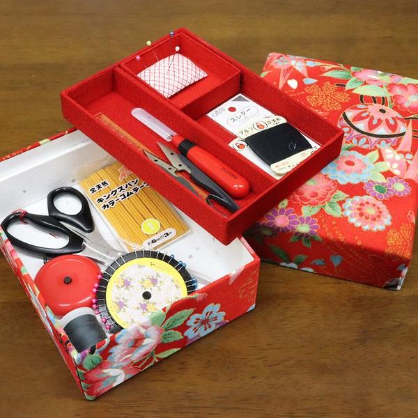 A red box with various sewing and crafting supplies, including scissors, pins, and other tools, is shown on a wooden surface.