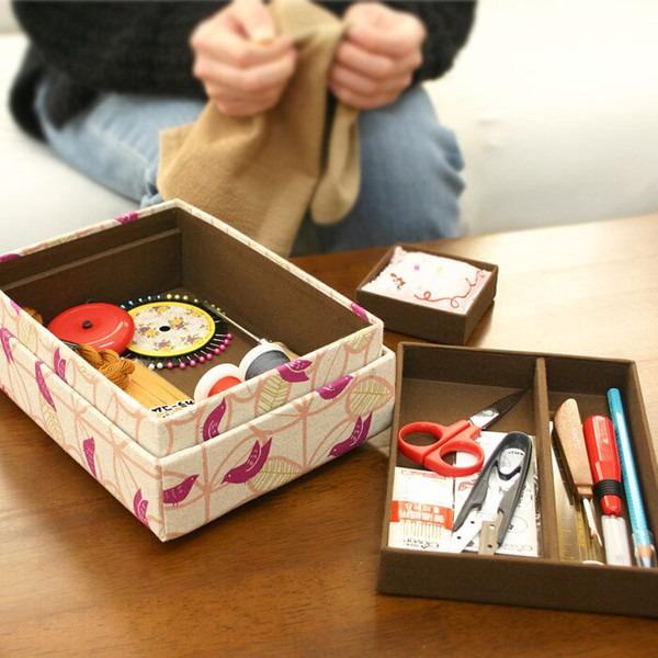 A person is sitting at a table, surrounded by various crafting supplies, including a box with sewing tools and a box with scissors and other office supplies.