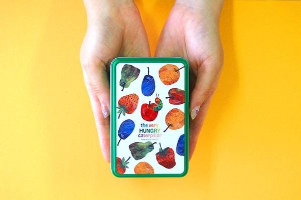 A pair of hands holding a small tin box with a colorful design featuring various fruits and vegetables, including apples, oranges, and bananas.