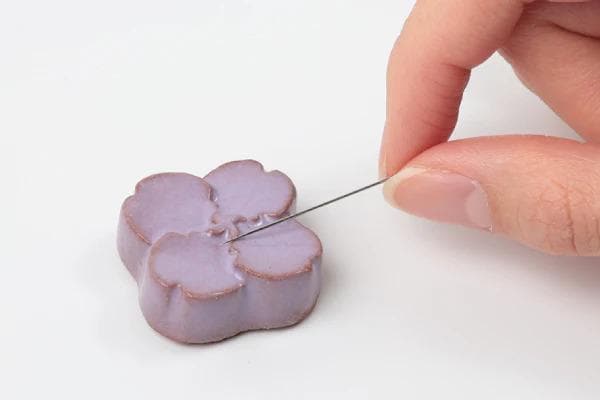 A hand is carefully threading a needle through a small purple flower-shaped object.