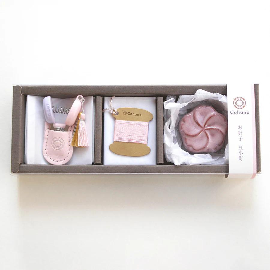 A set of three small items, including a pink keychain, a spool of thread, and a pink flower, arranged in a rectangular box.