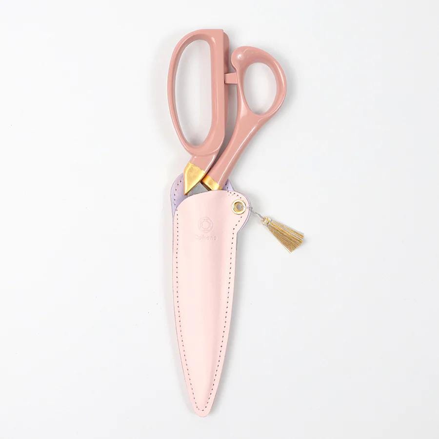 A pair of pink scissors with a tassel hanging from the handle, placed in a pink leather case against a white background.