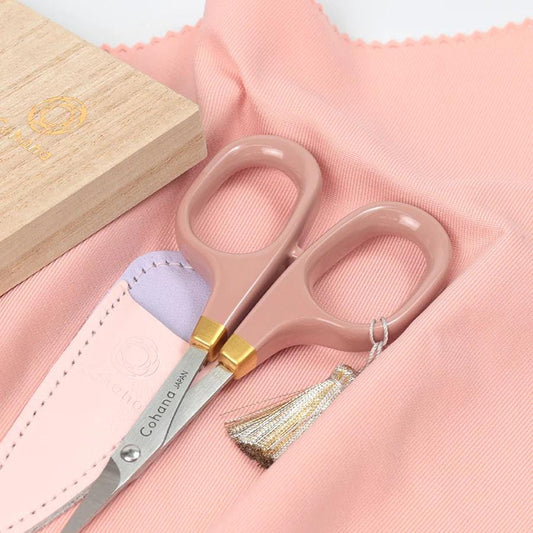 A pair of pink scissors with a tassel and a wooden box with a gold logo are placed on a pink fabric.
