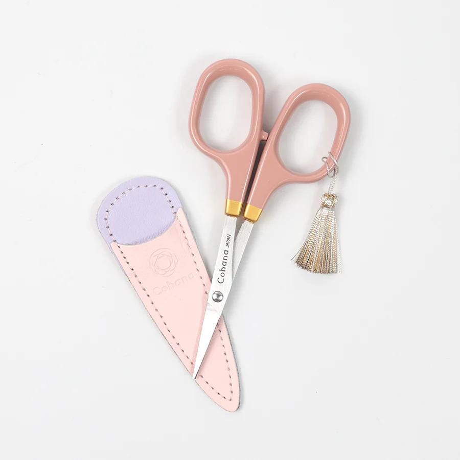 A pair of pink scissors with a tassel and a pink leather scissor case are placed on a white background.