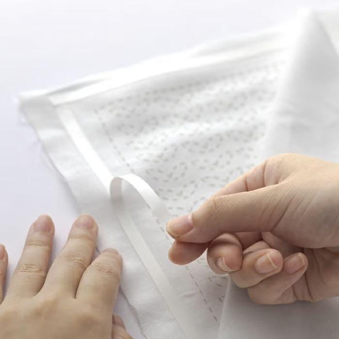 A close-up image of a person's hands holding a piece of white fabric with a patterned design, possibly sewing or crafting the fabric.