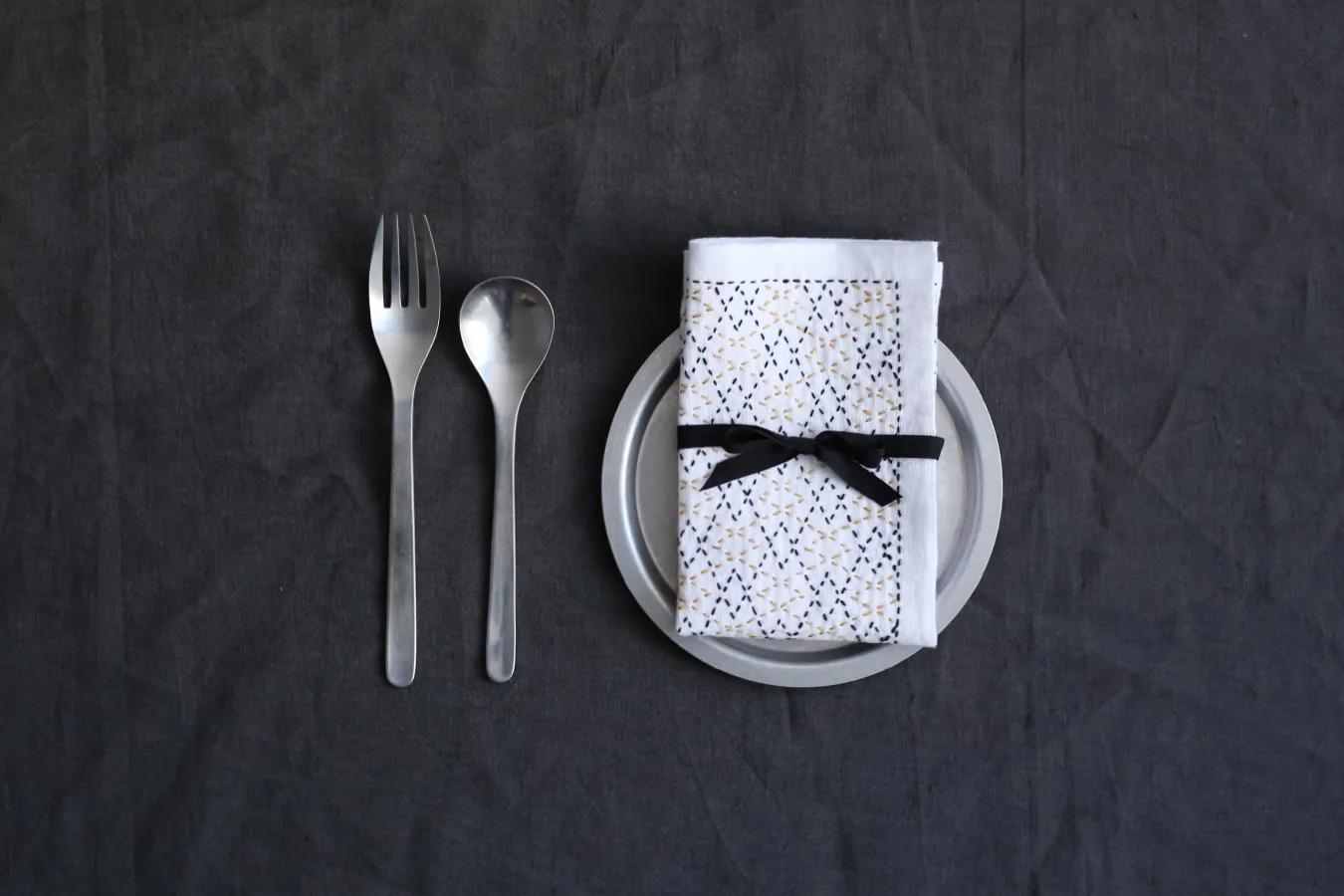 A set of silverware, including a fork, spoon, and knife, is arranged on a dark grey background, accompanied by a neatly folded napkin with a black ribbon tied around it.