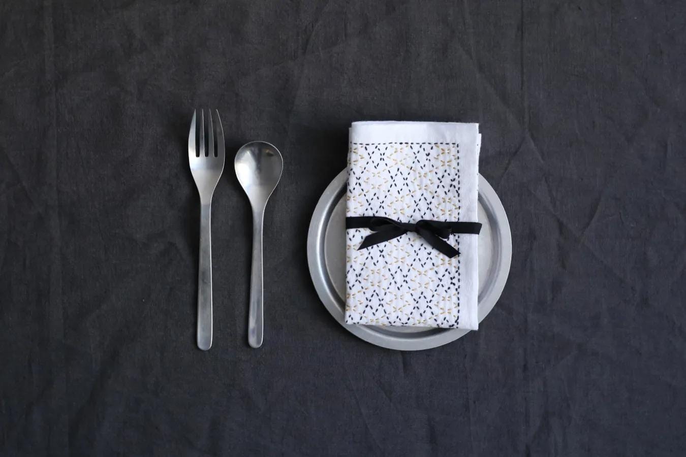 A set of silverware, including a fork, spoon, and knife, is arranged on a dark grey background, accompanied by a neatly folded napkin with a black ribbon tied around it.