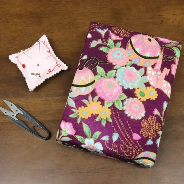 a pair of scissors, a small pink pin cushion, and a purple fabric with a floral pattern, all placed on a wooden surface.