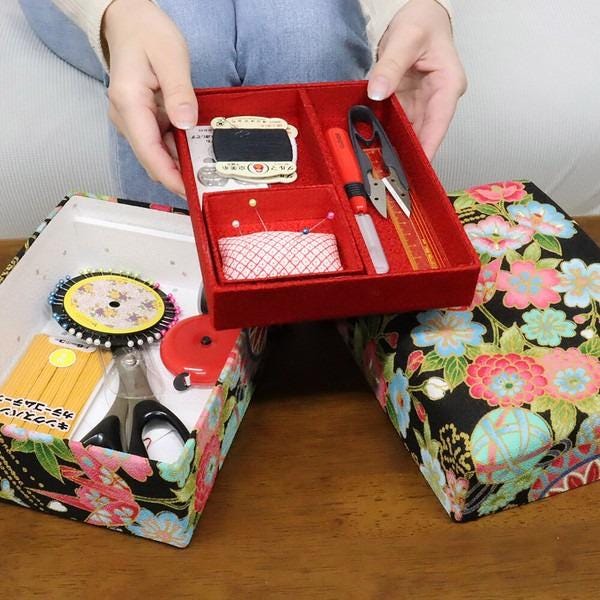 A person is holding a red box containing sewing supplies, including scissors, pins, and a sewing kit.