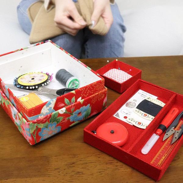 A person is sitting at a table, surrounded by various sewing and crafting supplies, including a red box with a floral pattern, a red tray with sewing tools, and a small red box with a white label.