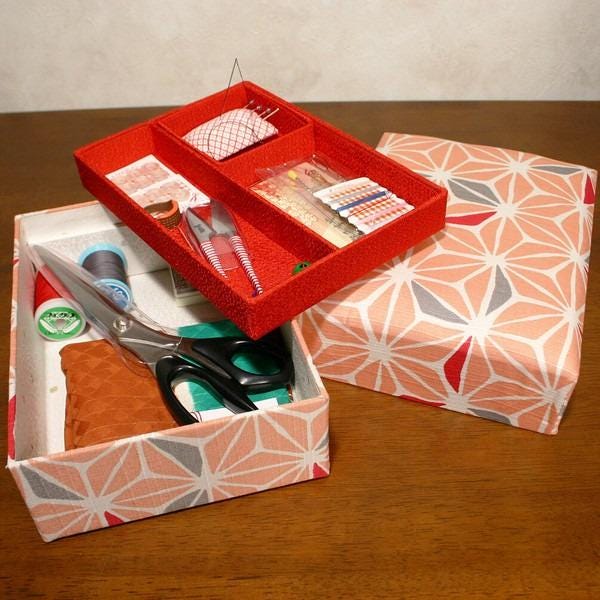 A red storage box with compartments containing sewing supplies, including scissors, pins, and needles, sits on a wooden surface.
