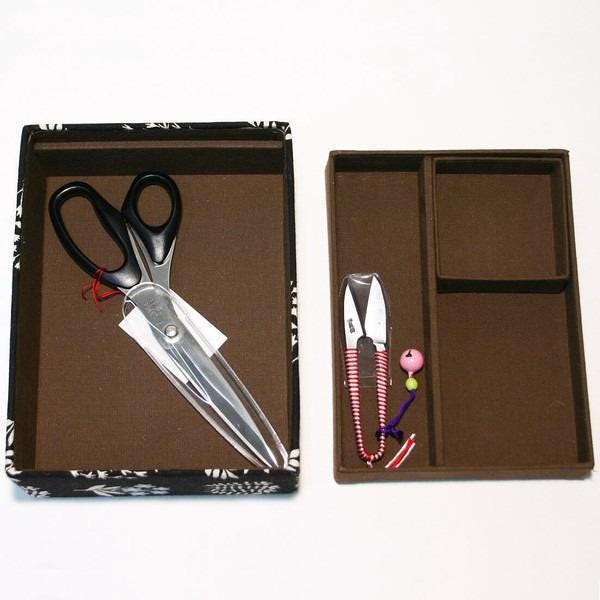 a pair of scissors and a pair of scissors with a pink handle, both placed inside a brown box with compartments.