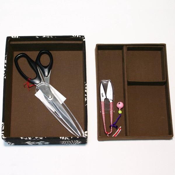 a pair of scissors and a pair of scissors with a pink handle, both placed inside a brown box with compartments.