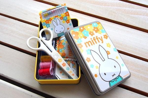 A yellow box containing a pair of scissors, a sewing kit, and a small tin with a bunny design, all placed on a wooden surface.