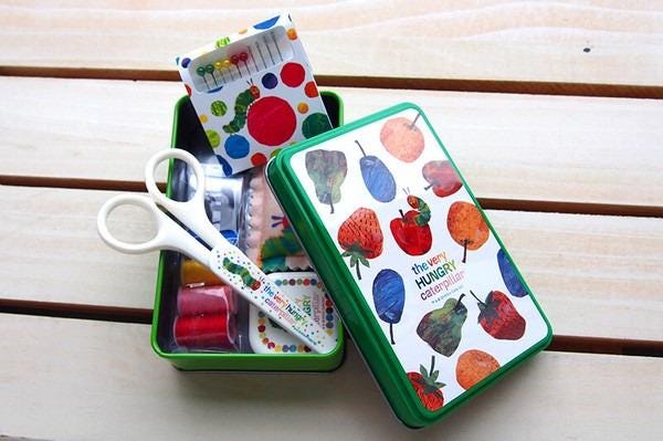 A green tin box with a pattern of colorful fruits and vegetables, including strawberries, pears, and oranges, along with a pair of scissors.
