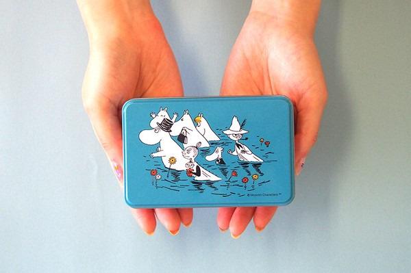 A pair of hands holding a blue tin with a cartoon illustration of a group of characters, including a hippopotamus, on a body of water.