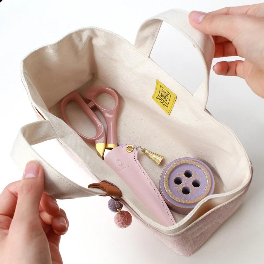 A hand is holding a small white tote bag with various sewing and crafting supplies inside, including pink scissors, a purple button, and a pink pouch.