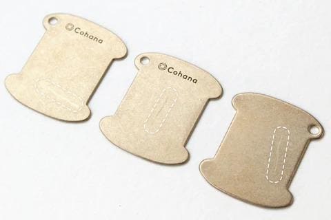 Three identical metal spool-shaped tags with the brand name "Cohana" printed on them.