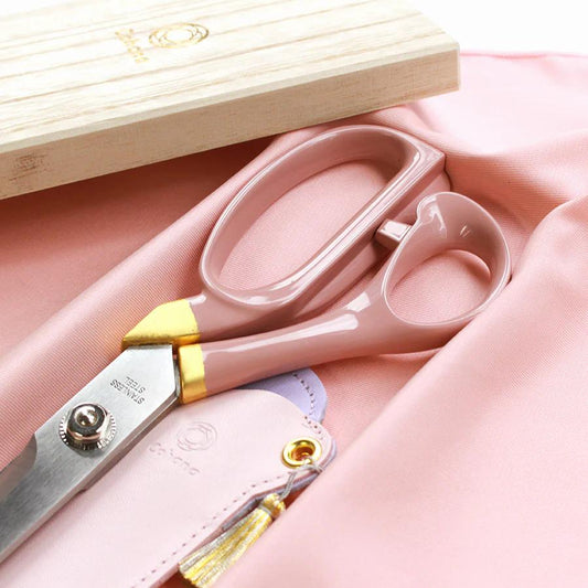 A pair of pink scissors with gold accents and a wooden box with a gold logo are displayed on a pink fabric surface.