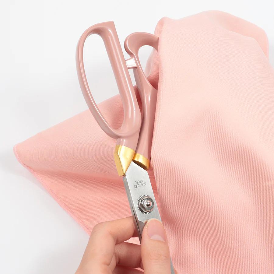 A hand holding a pair of pink scissors with a pink fabric in the background.