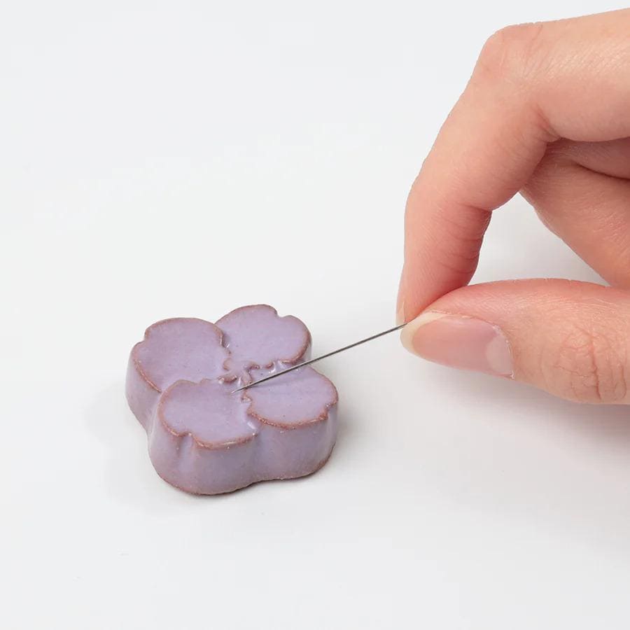 A hand is holding a needle and thread, preparing to sew a purple flower-shaped object.