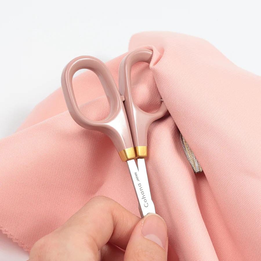 A hand holding a pair of pink scissors with the blades open, resting on a pink fabric.