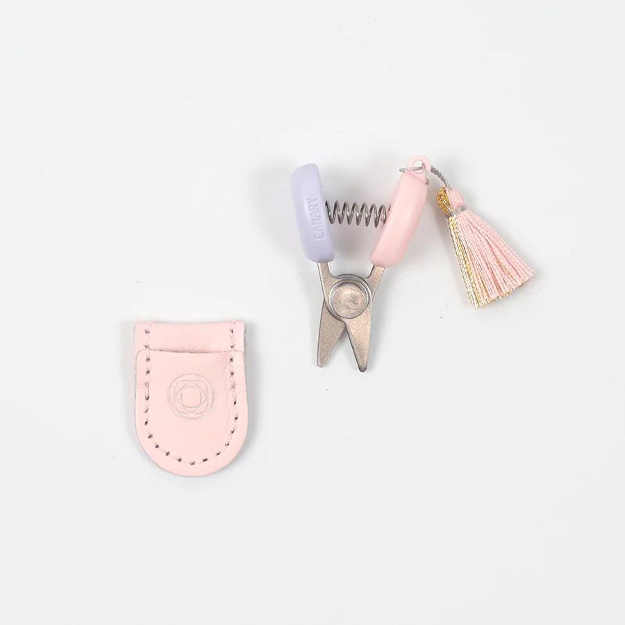 A pair of scissors with a pink handle and a tassel, and a small leather pouch with a circular logo on it, all placed on a white background.