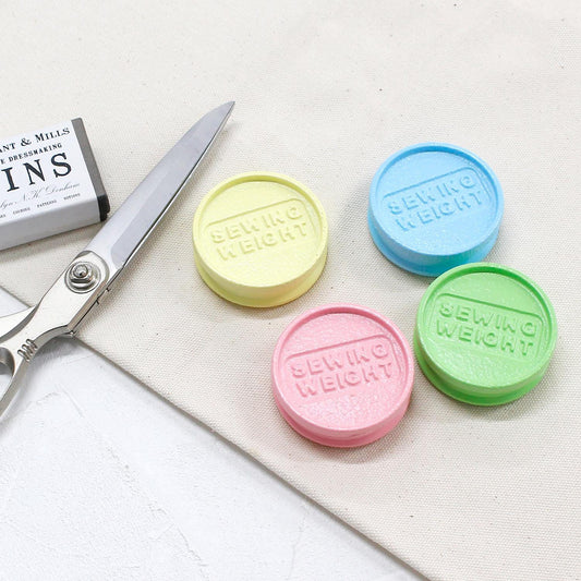 a pair of scissors, four colorful sewing weight lids, and a piece of fabric on a white surface.