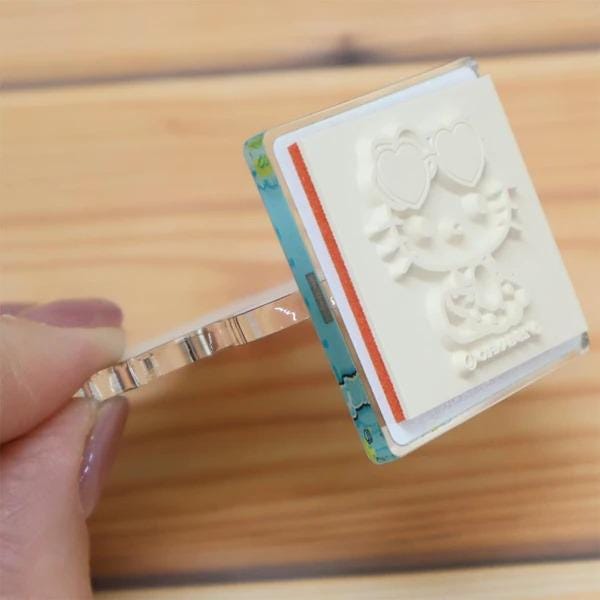 A hand is holding a white stamp with a cartoon character design on it, which appears to be a playful and creative item.