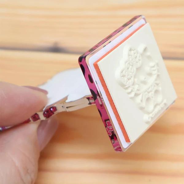 A hand is holding a white stamp with a pink and black design on it, which appears to be a decorative item.