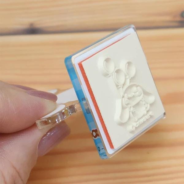 A hand is holding a white stamp with a cartoon character on it, which is placed on a wooden surface.
