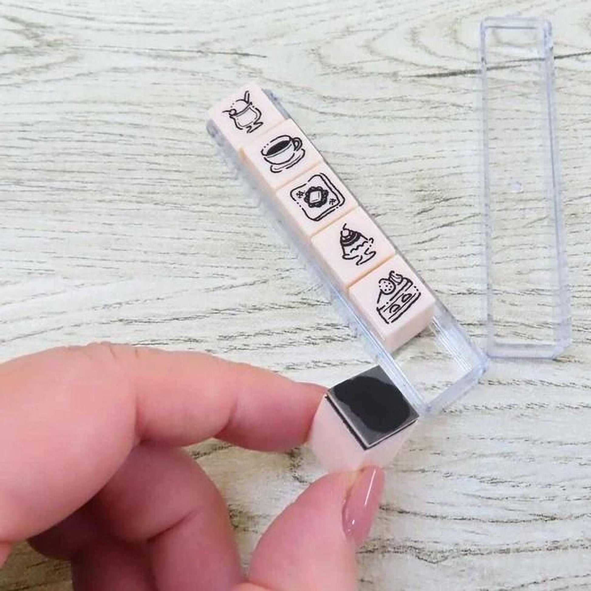 A hand is holding a clear plastic container with five small, square-shaped objects with black and white illustrations on them.