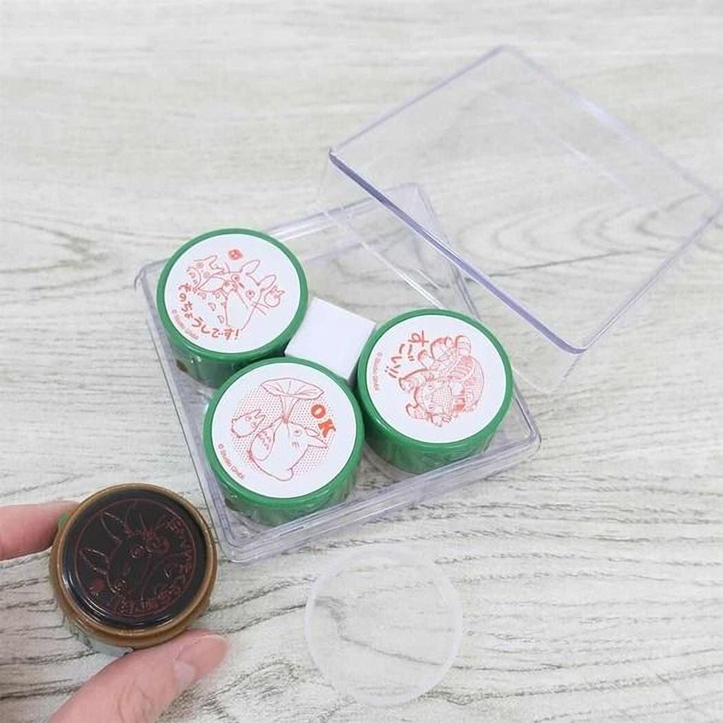 a person holding a small green and white stamp with a red and white design, placed on a white surface next to a clear plastic container with a lid.