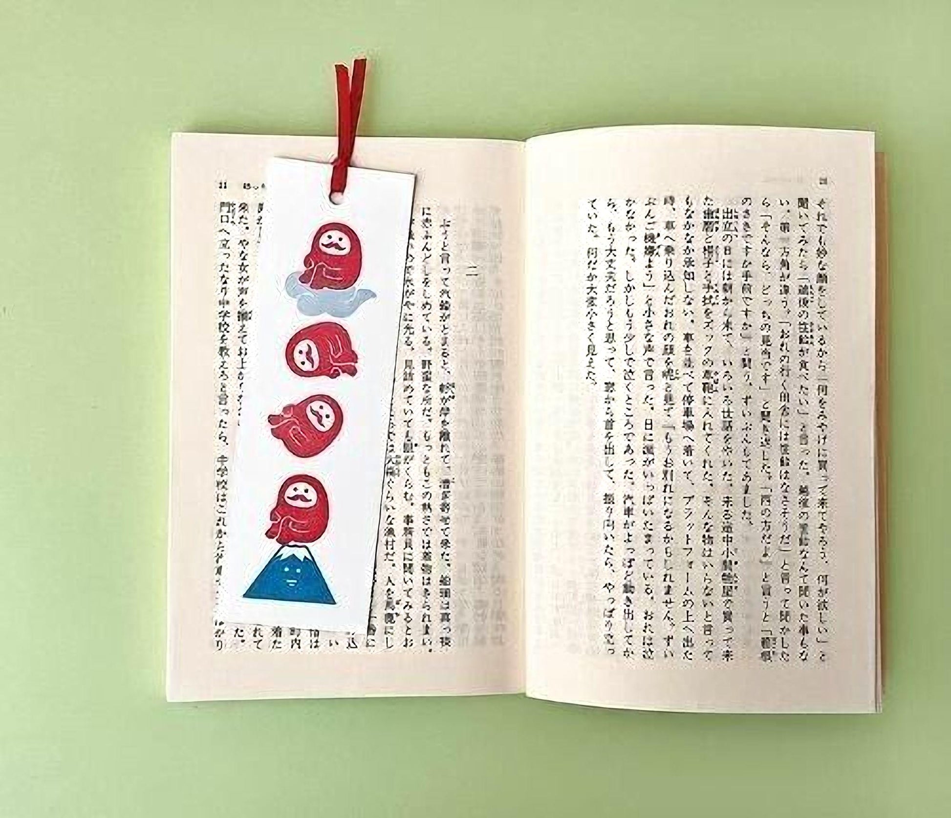 An open book with Japanese text on the pages, and a red bookmark with a cartoon character design on the left page.