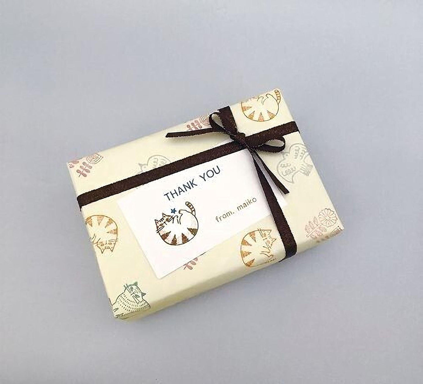 A gift box wrapped in brown paper with a bow, featuring a "Thank You" card with a cat illustration and the text "from makoto" printed on it.