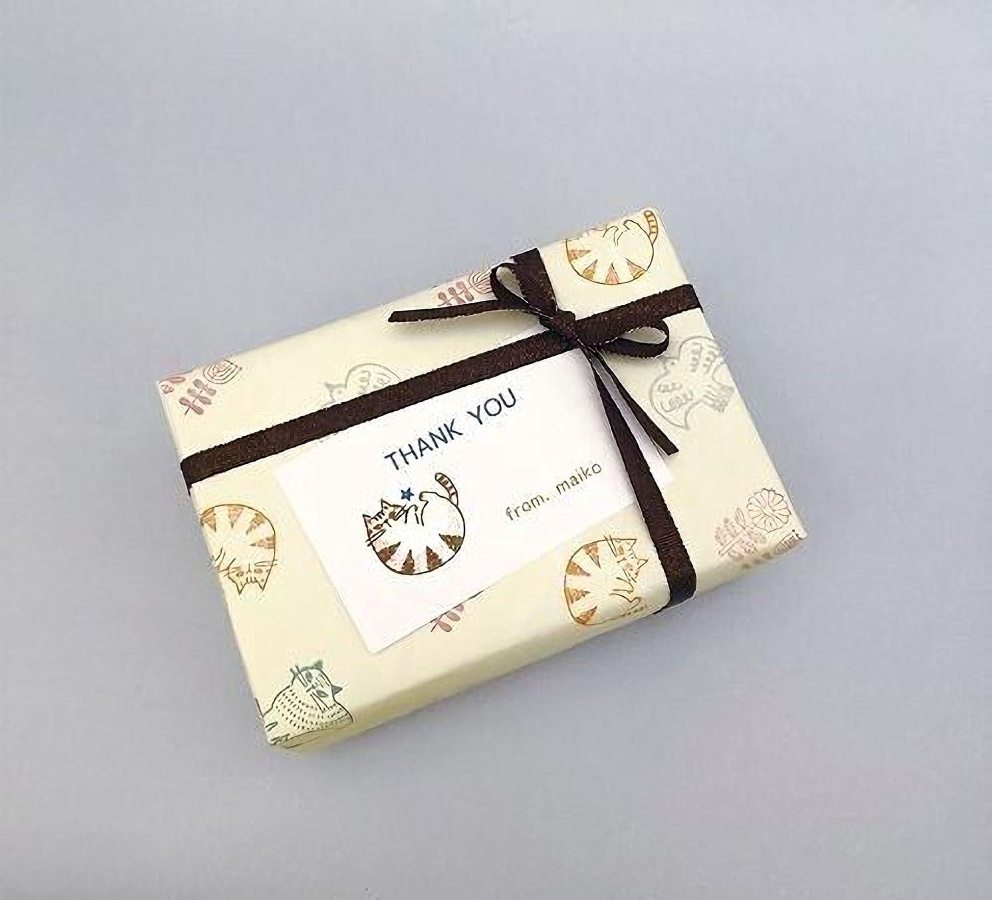 A gift box wrapped in brown paper with a bow, featuring a "Thank You" card with a cat illustration and the text "from makoto" printed on it.