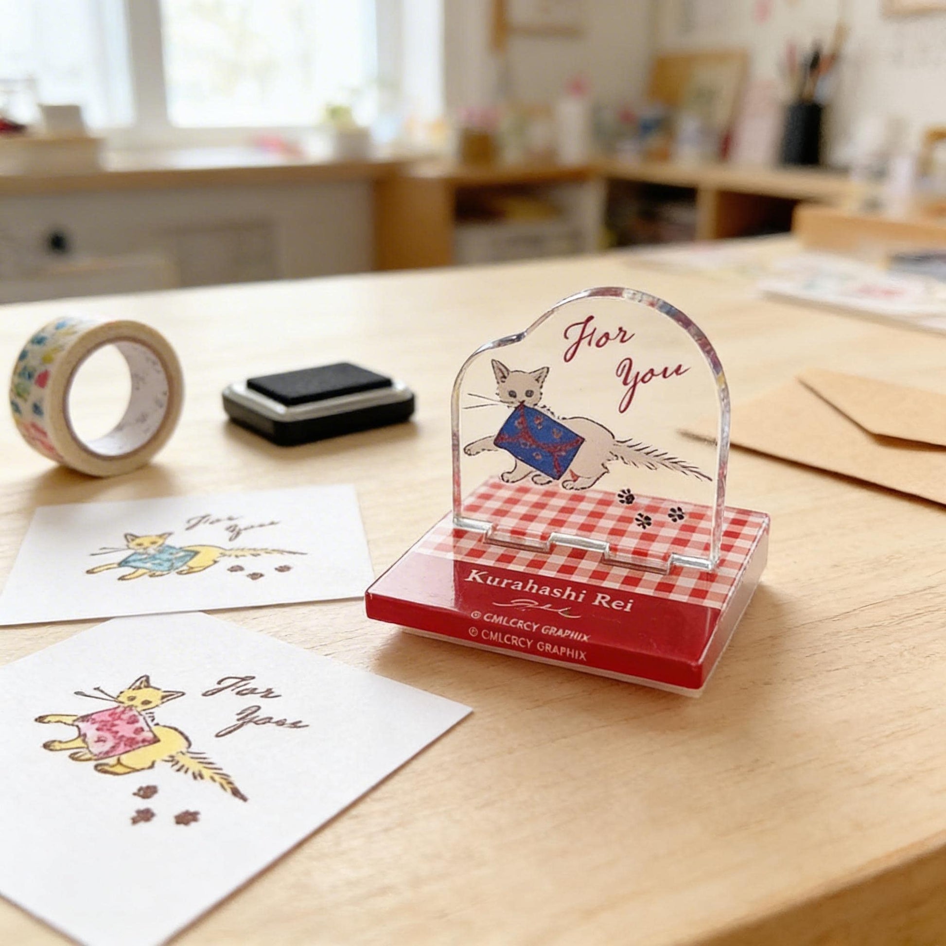 a red and white checkerboard-patterned stamp with a cat illustration, placed on a wooden table. There are also two pieces of paper with cat illustrations and the text "For You" written on them.