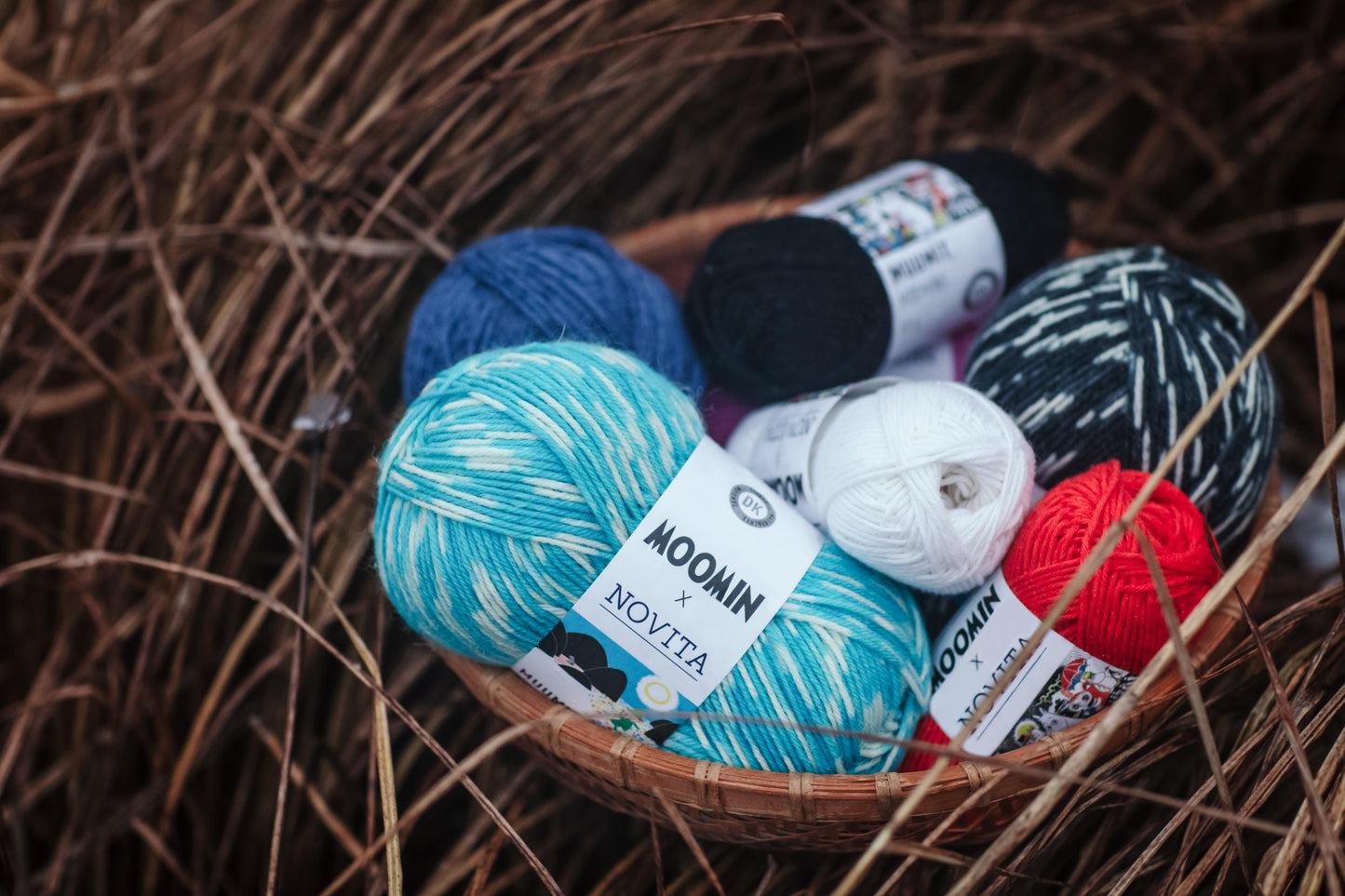 A basket filled with various balls of yarn, including blue, black, white, and red yarn, resting on a bed of straw or grass.