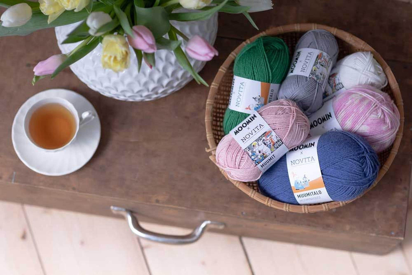 A wooden table with a white vase holding pink and white tulips, a basket of yarn with various colors, and a cup of tea.