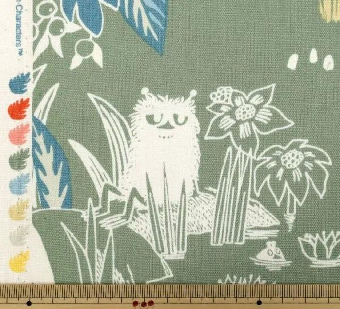 a close-up view of a fabric with a whimsical, cartoonish design. The design includes various elements such as a cat, flowers, leaves, and other abstract shapes. The fabric appears to be a piece of cloth or textile, with a ruler visible in the bottom right corner.