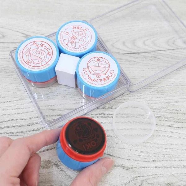 A hand is holding a blue and red stamp in front of a clear plastic container with four other similar stamps inside.