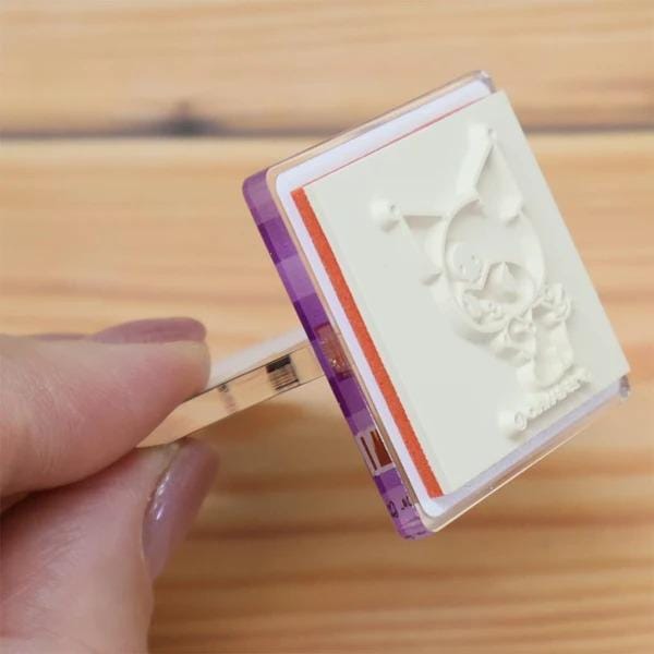 A hand is holding a white stamp with a cartoon character on it, which is placed on a wooden surface.