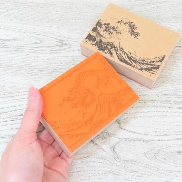 A hand is holding a rectangular stamp with an orange background and a black wave design on it, placed on a wooden surface.