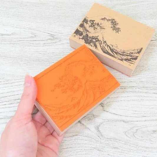 A hand is holding a rectangular stamp with an orange background and a black wave design on it, placed on a wooden surface.