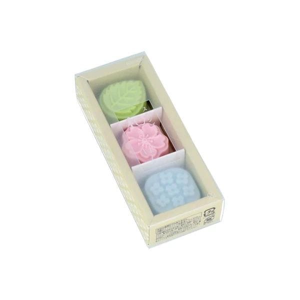 A rectangular box containing three small, colorful objects, likely a set of three different colored candles or small decorative items.