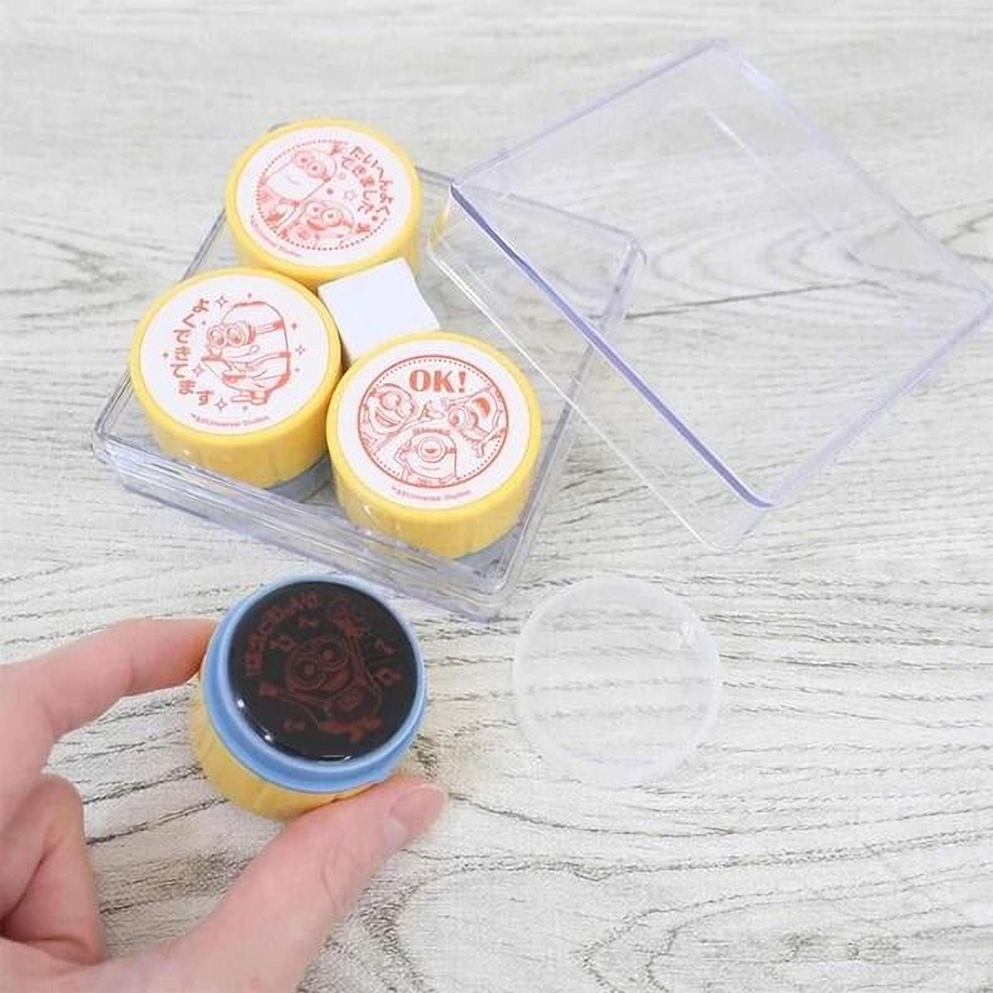 a hand holding a stamp with a cartoon character on it, along with a clear plastic container and a blue stamp pad.