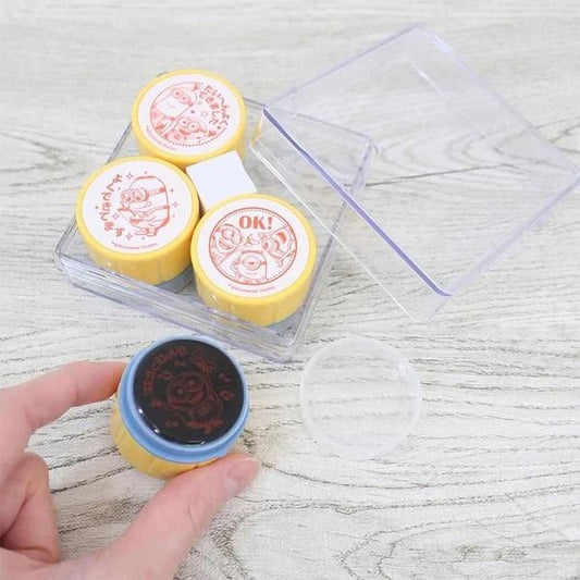 a hand holding a stamp with a cartoon character on it, along with a clear plastic container and a blue stamp pad.