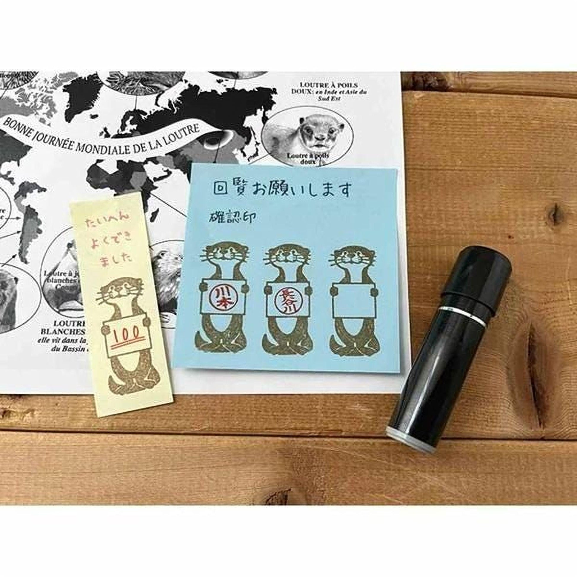 a blue sticky note with a drawing of four otters holding signs, a yellow sticky note, and a black marker on a wooden surface.