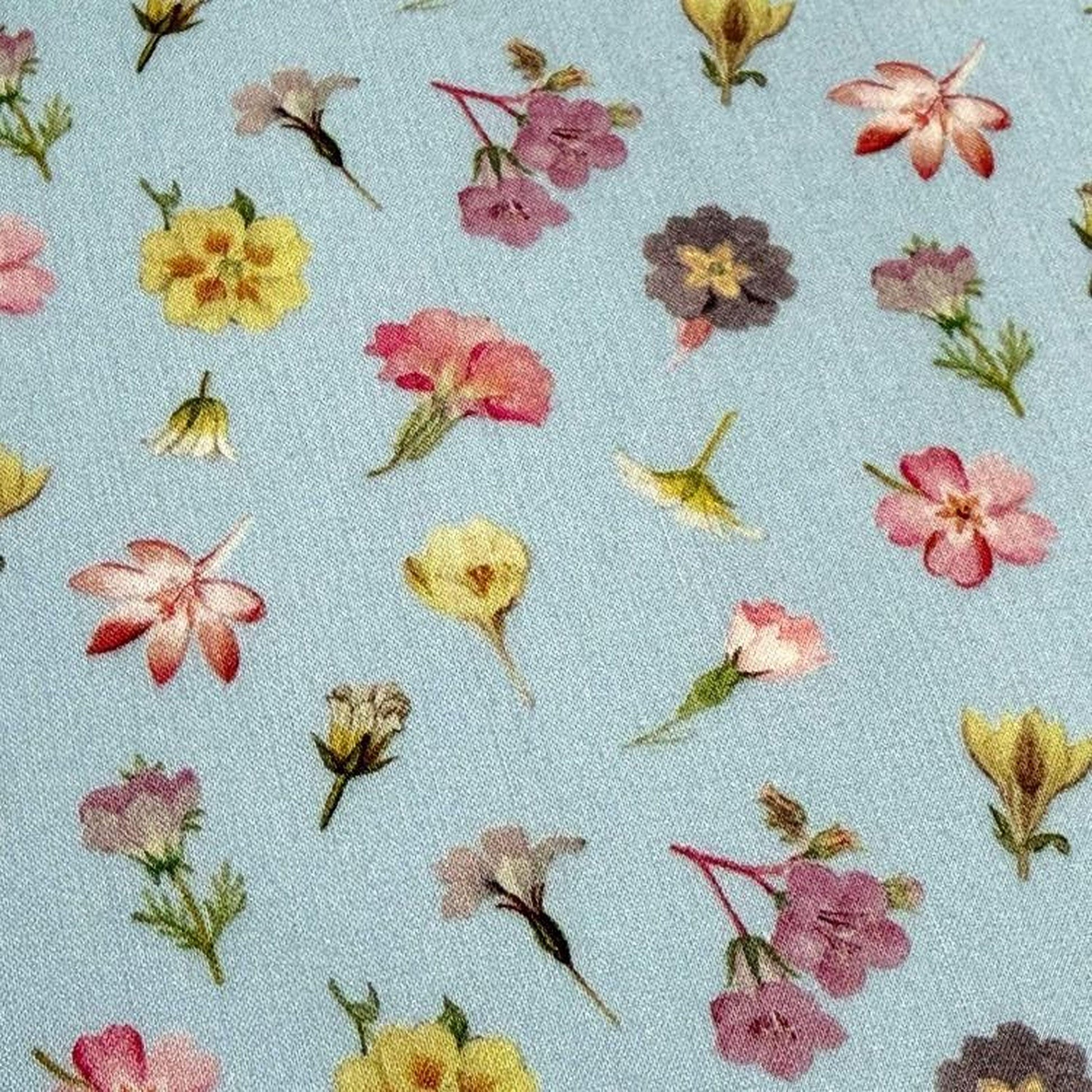 a close-up view of a fabric with a repeating pattern of various colorful flowers, including yellow, pink, and purple flowers, against a light blue background.